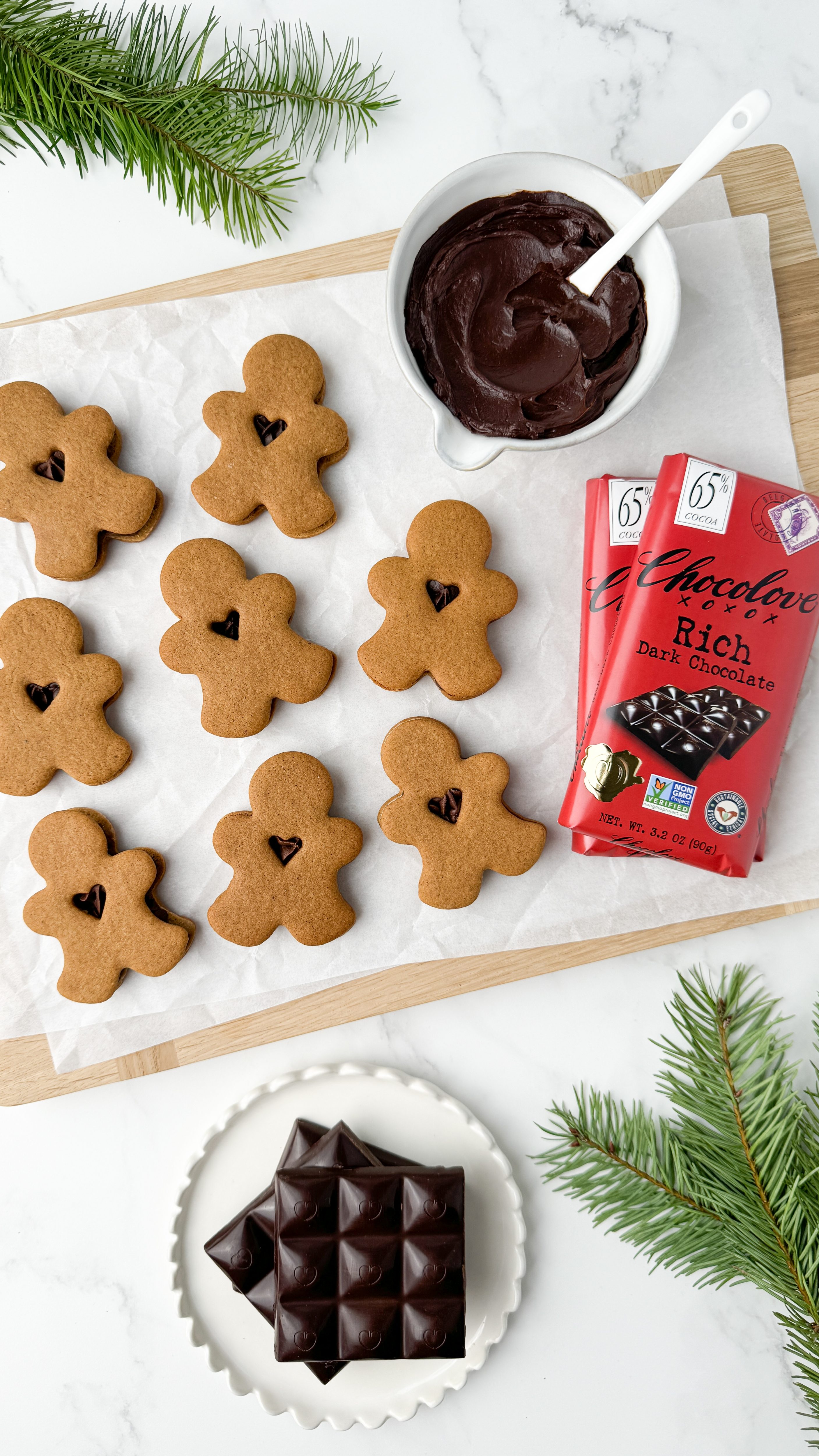 Festive gingerbread sandwich cookies with small heart cutouts, filled with rich dark chocolate ganache, arranged on parchment paper. A bowl of smooth ganache with a white spoon sits nearby, along with two Chocolove Rich Dark Chocolate Bars and a plate of dark chocolate squares, surrounded by fresh evergreen sprigs